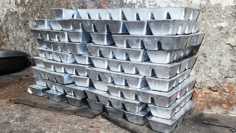 zinc and aluminum ingot stacks rest on the ground before a rough, weathered wall.