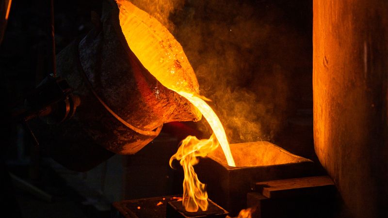 Se vierte un metal fundido de color naranja brillante desde un crisol a un molde; se ven llamas.